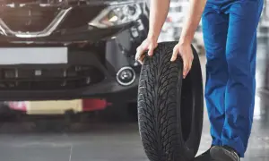 Mechanik w niebieskich spodniach pokazuje, jak założyć opony w dobrym kierunku, trzymając nową oponę przed czarnym samochodem.