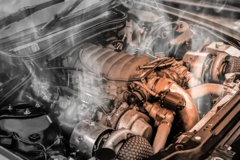 Dym unosi się z silnika samochodu, wskazując na jednorazowe przegrzanie. Widoczne są turbosprężarki i skomplikowane przewody.