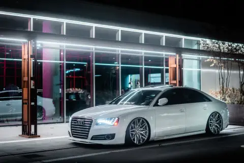 Białe Audi z obniżonym zawieszeniem **air ride** stoi przed nowoczesnym budynkiem z neonowym oświetleniem.