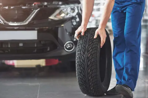 Mechanik w niebieskich spodniach pokazuje, jak założyć opony w dobrym kierunku, trzymając nową oponę przed czarnym samochodem.