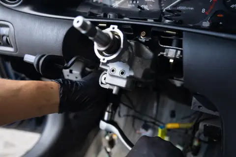 Mechanik w czarnej rękawiczce dokręca śrubę kluczem nasadowym przy kolumnie kierownicy.