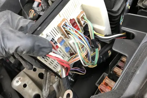 Ręka w rękawiczce manipuluje skrzynką z bezpiecznikami w Renault Scenic 2. Widać kolorowe przewody i oznaczenia bezpieczników.